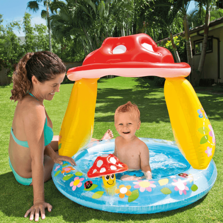 A happy toddler splashing in the Intex Mushroom baby pool with canopy outdoors on the grass while his mother watches.